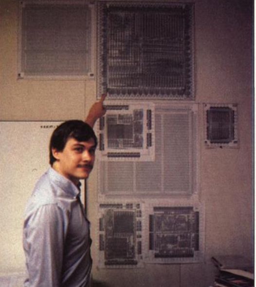Duck Renn pointing to the large chip layout diagrams on the laboratory wall